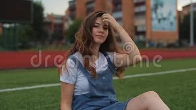 一幅美丽的黑发女人的肖像，穿着蓝色的阳光，坐在体育场的一个足球球门附近的草坪上。 阳光照射